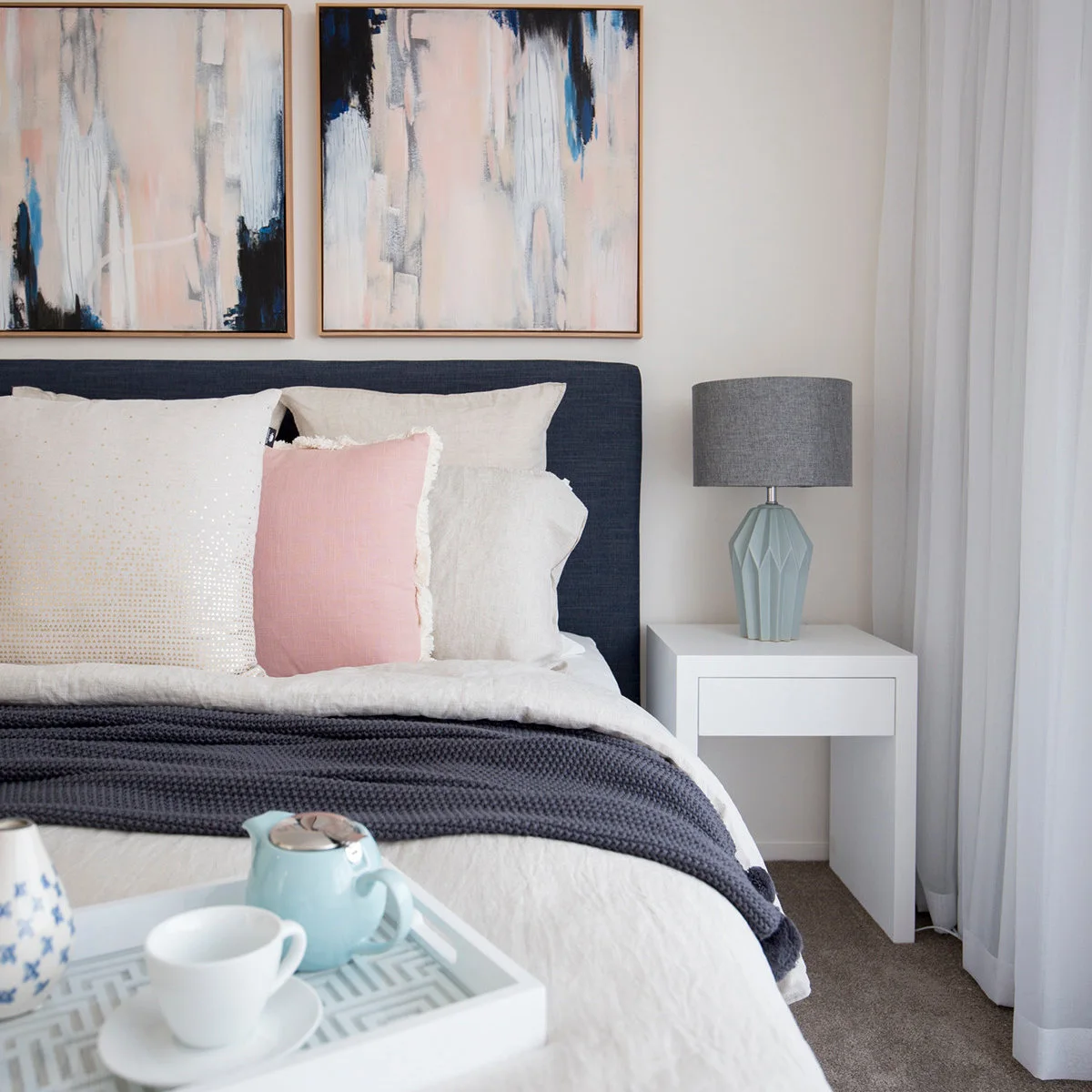 Two pale pink and blue abstract paintings above a navy headboard beside a white table and blue-gray lamp.