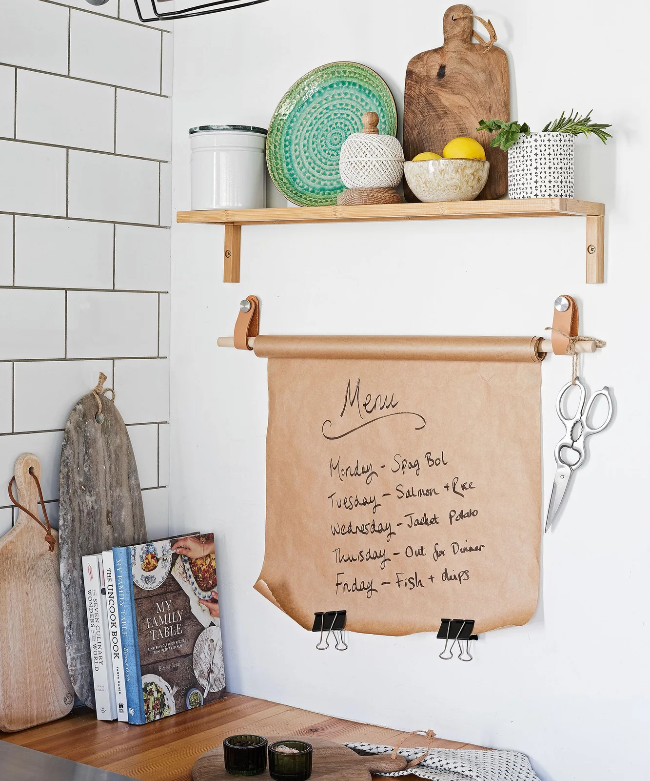 Wall-mounted brown paper menu roll below a small kitchen shelf with a green plate and a wooden board.