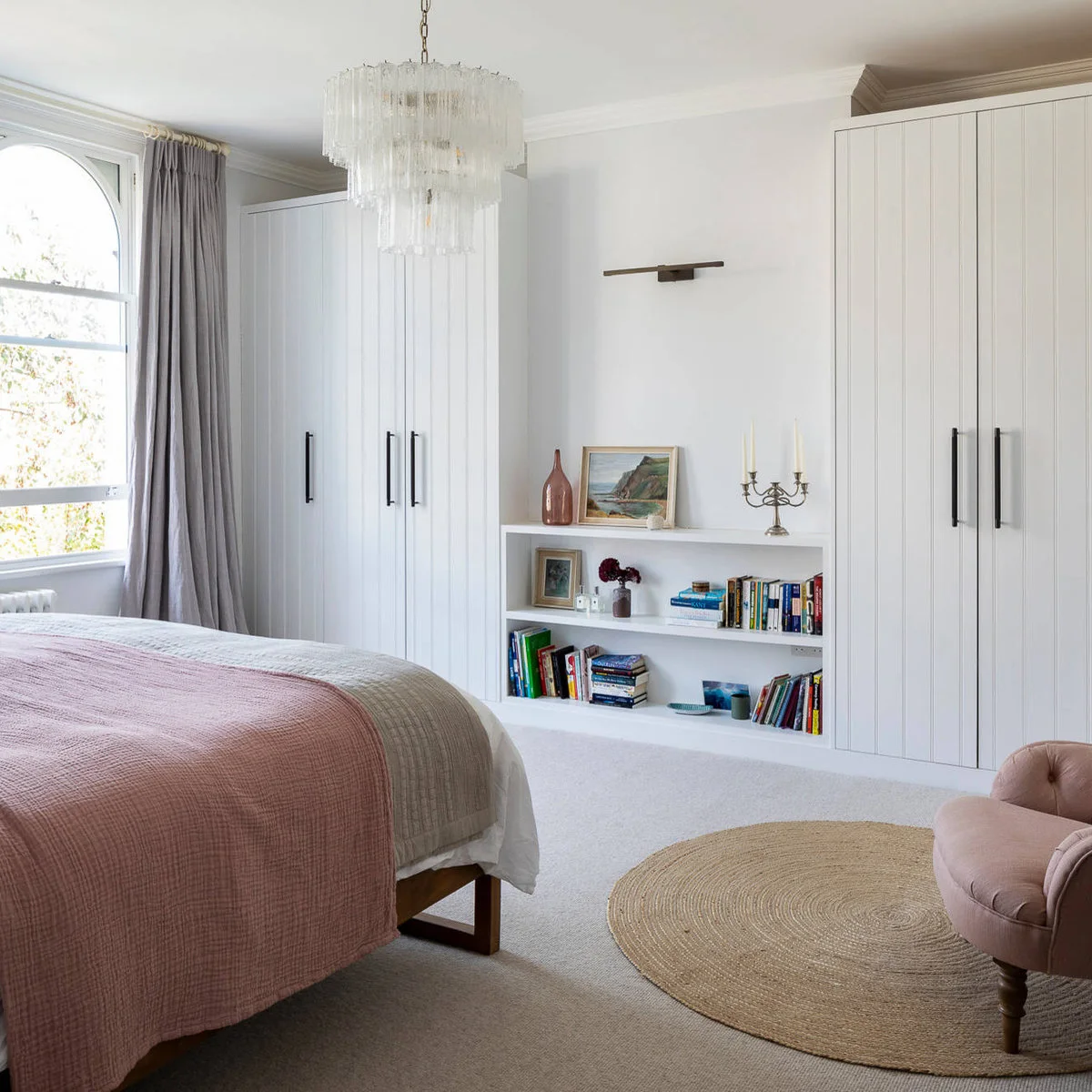 Pale bedroom with a blush bed cover, small pink chair and white built-in wardrobes