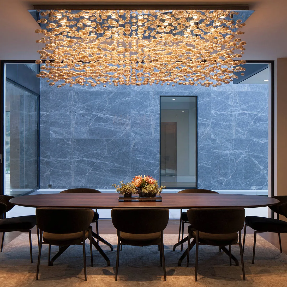 Long dark oval dining table with dark chairs beneath a broad glowing decorative chandelier