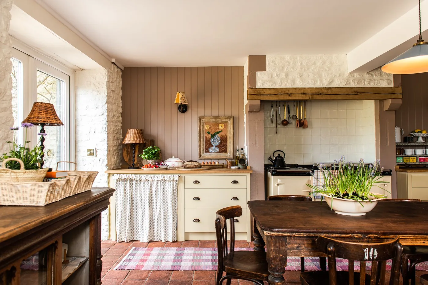 Cottage kitchen with pale cupboards, a light fabric skirt, pink lower runner, wooden table and painted wall paneling beside a cooking alcove.