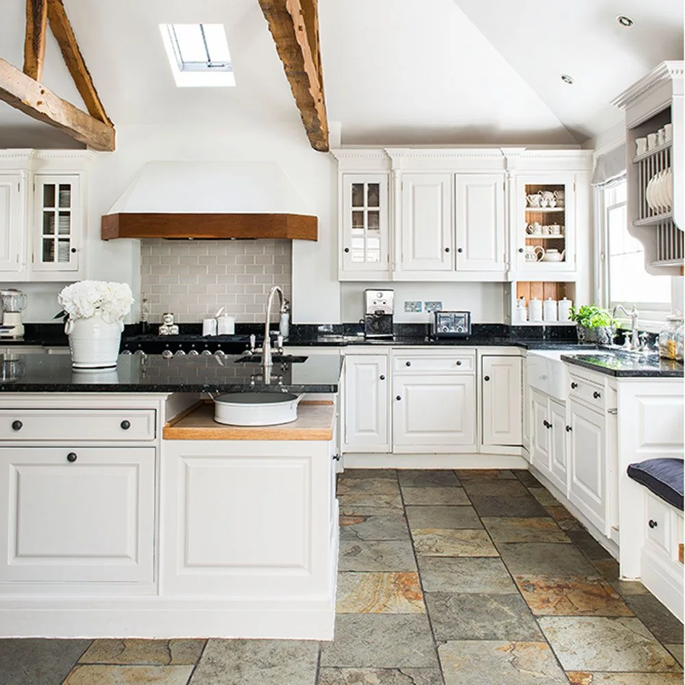 White framed kitchen cupboards and glazed doors sit below wood beams beside dark counters, a pale tiled cooking wall and warmer inset storage surfaces