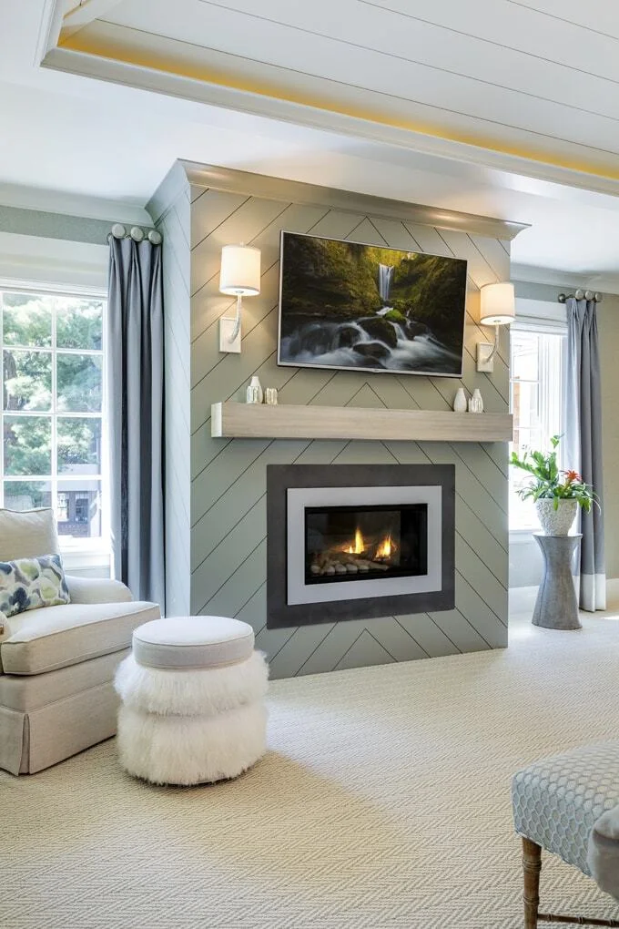 A gray-green fireplace wall has diagonal chevron boards, a pale mantel, paired shaded wall lights and a television above a dark-bordered fire opening.