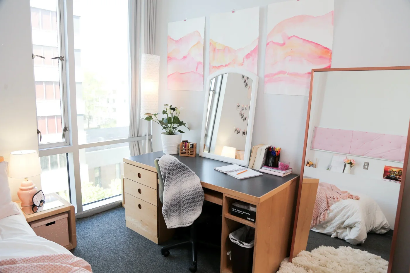 Wood dorm desk with mirror, notebooks, small stationery holder and plant beside tall windows