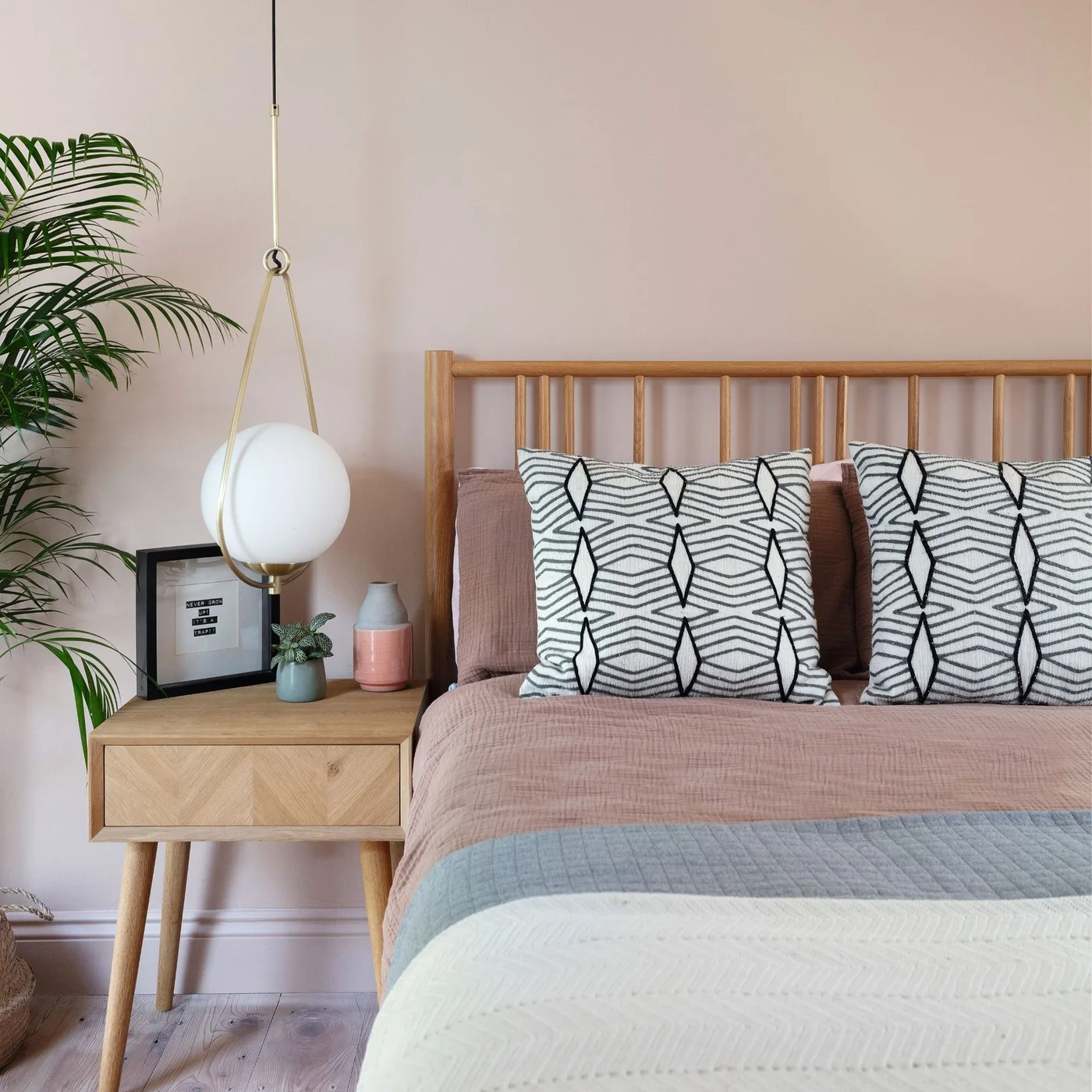 A hanging white globe light sits beside a wood spindle headboard and a small wood drawer table against a pale pink wall with patterned pillows.