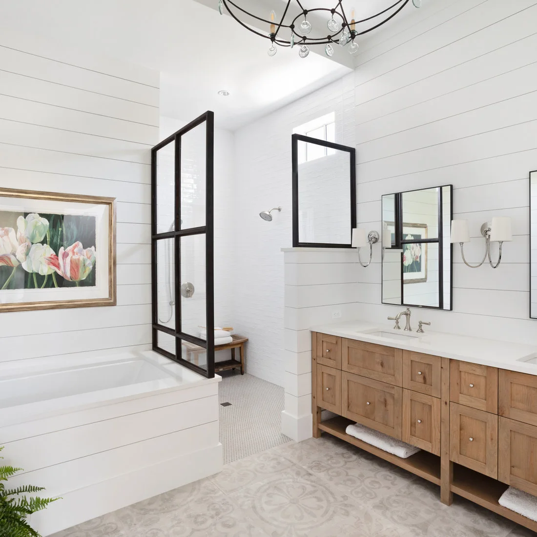 Pale small mosaic tiles inside a black-framed shower meet larger patterned bathroom floor tiles beside a timber vanity.