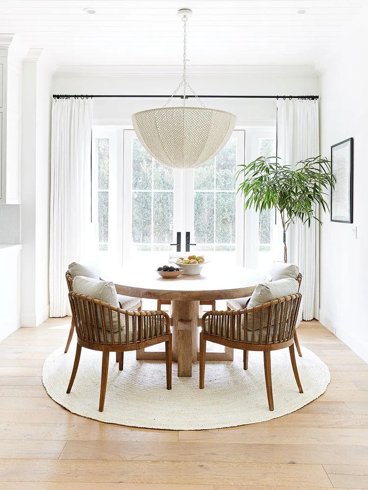 Dining corner with round wooden table, woven-backed chairs, woven pendant, pale round rug and white walls.