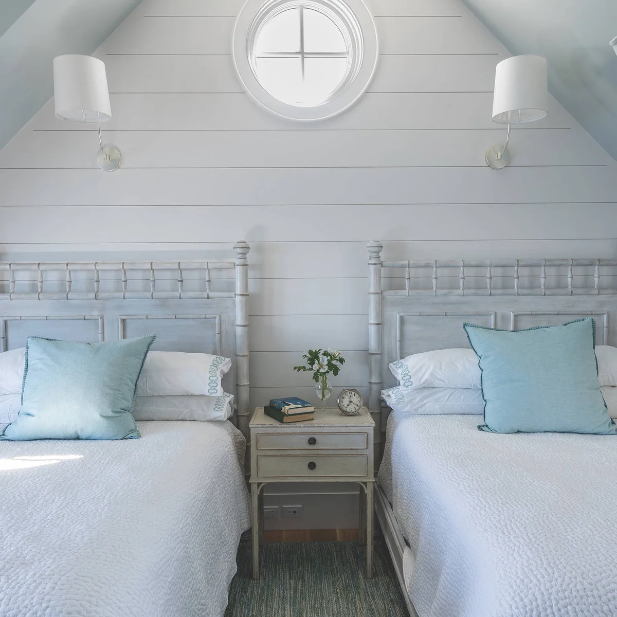 Two pale beds with aqua cushions, a narrow two-drawer table and wall lamps beneath a round window