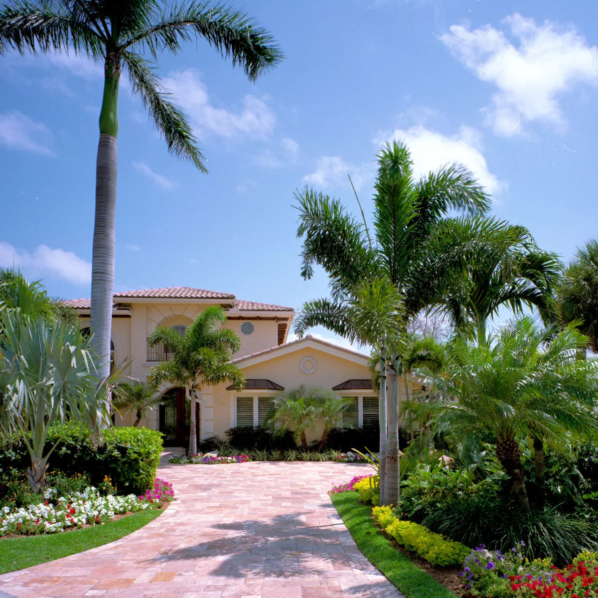 A broad pink-toned paved drive with palms, low colorful planting and a cream house visible beyond.