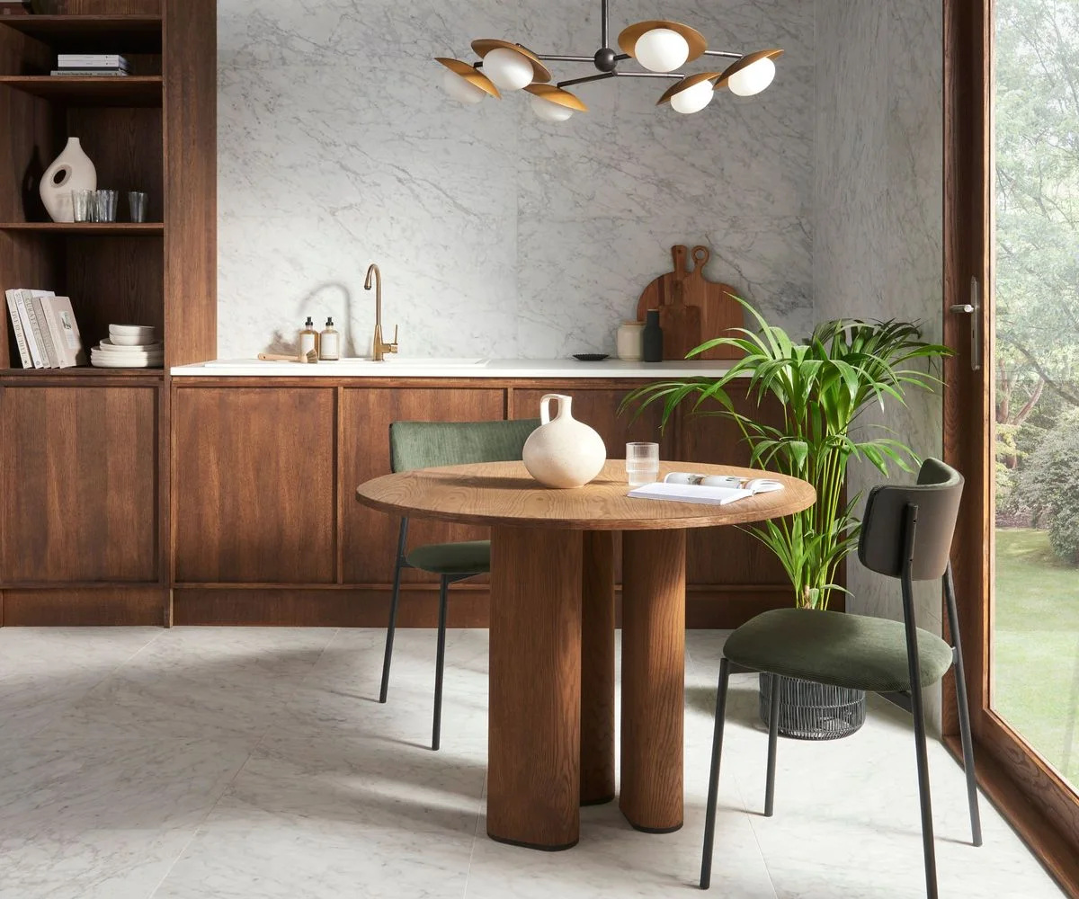 A round wooden table and muted green chairs stand beside wooden kitchen cabinets, a pale veined backsplash, open shelves and broad glazing