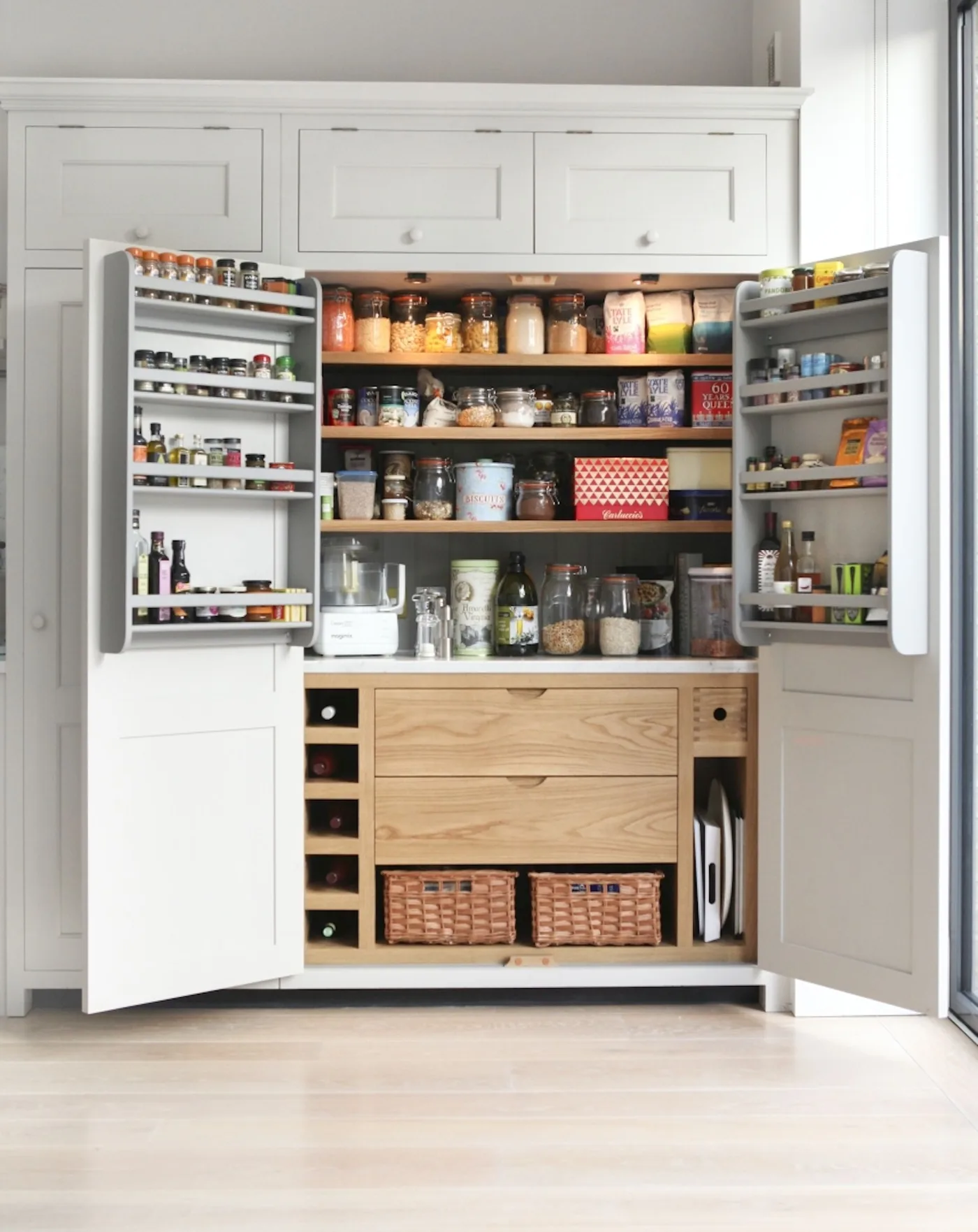 Country kitchen pantry cabinet with open pale doors, wooden shelves, jars, door-mounted spice racks, lower drawers, baskets and bottle cubbies.
