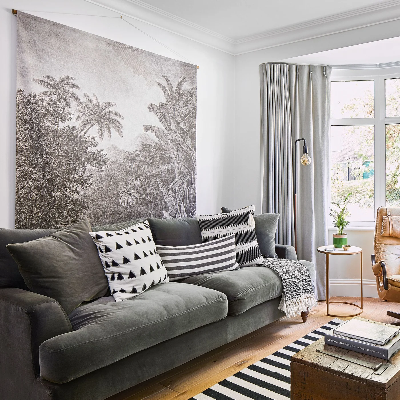 A large monochrome botanical wall hanging sits above a gray sofa with striped cushions beside a bright window