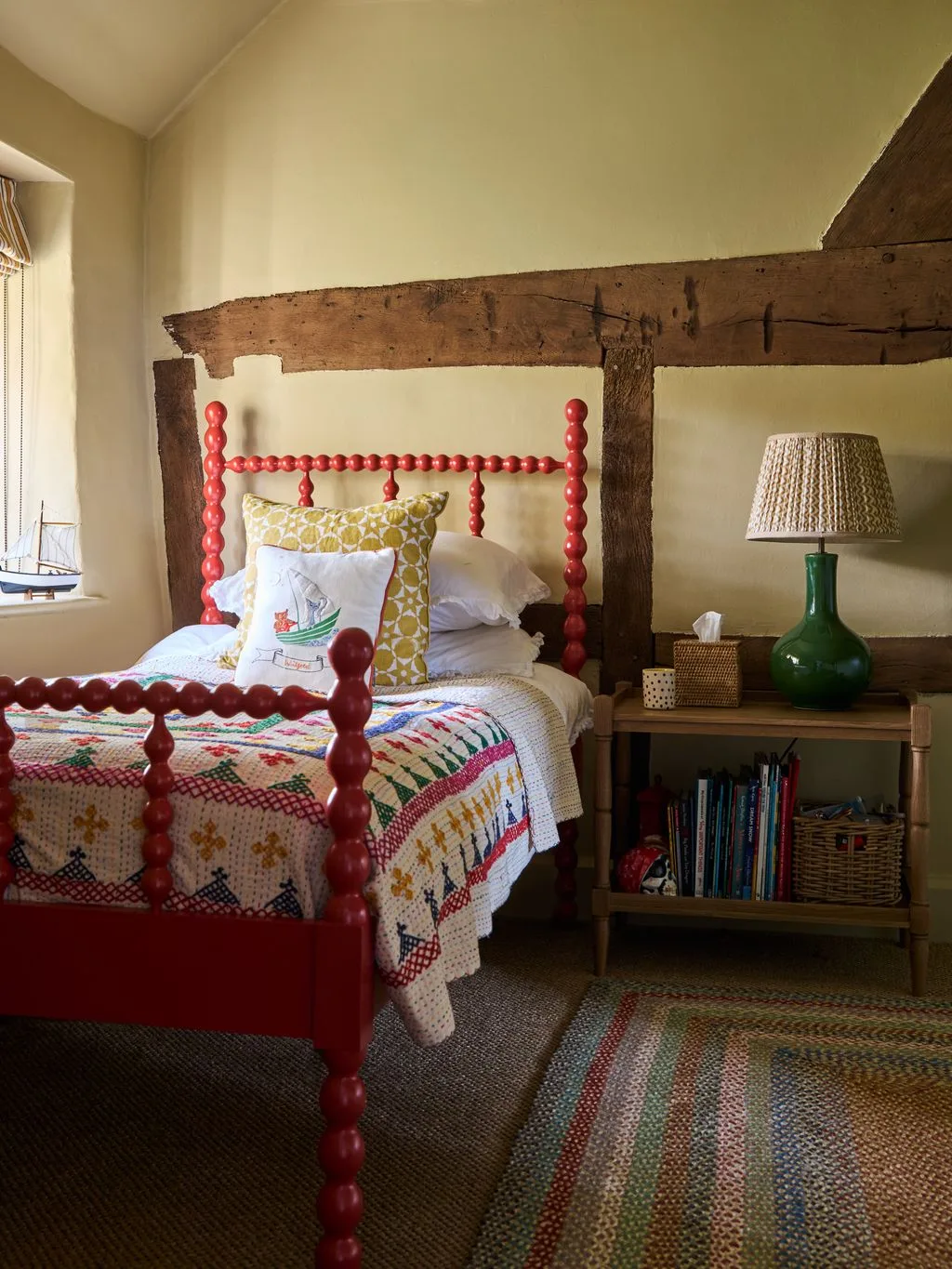 Single cottage bedroom with red turned bed frame, exposed wall beam, patchwork quilt, yellow cushion, green lamp and small book shelf