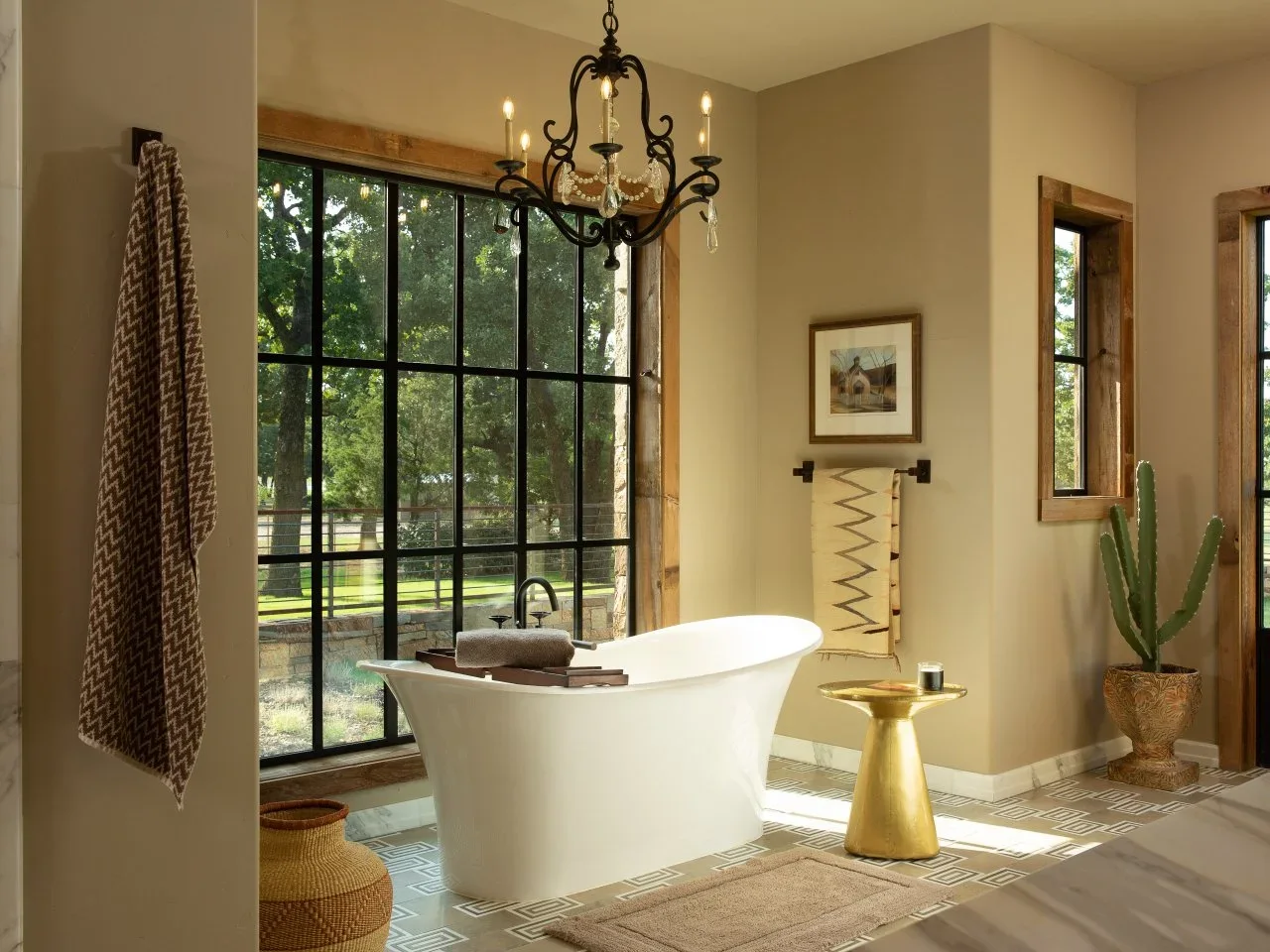 A cactus in a decorative pot stands near a white tub, timber-framed windows and pale warm walls.