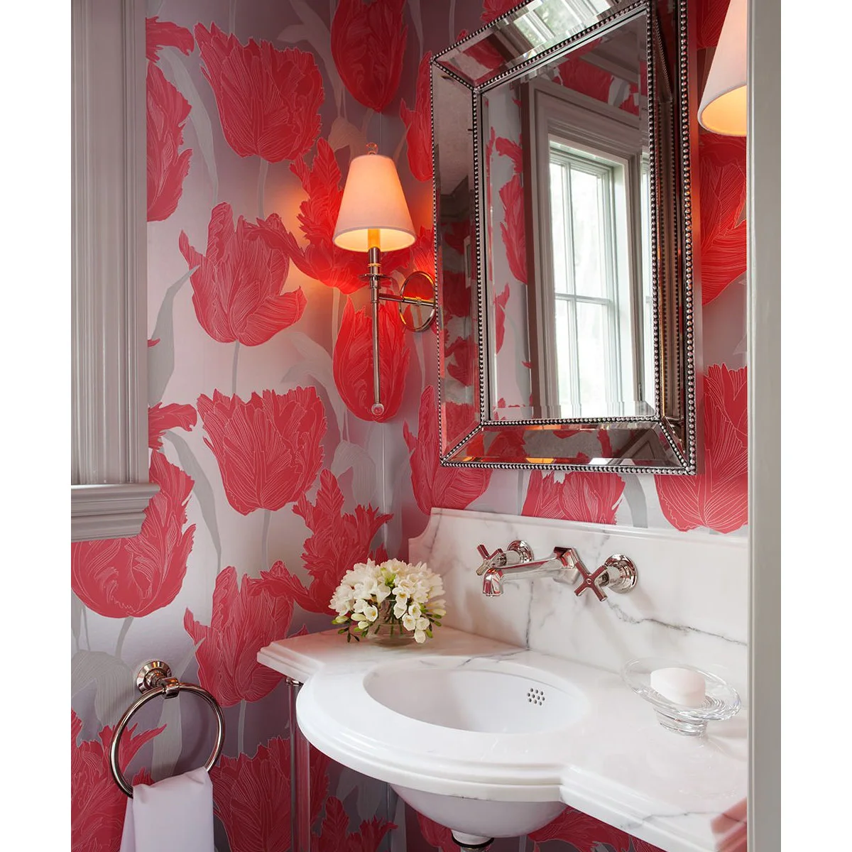 Large pink floral wallpaper around a curved white sink, with a mirrored frame and a shaded wall light.