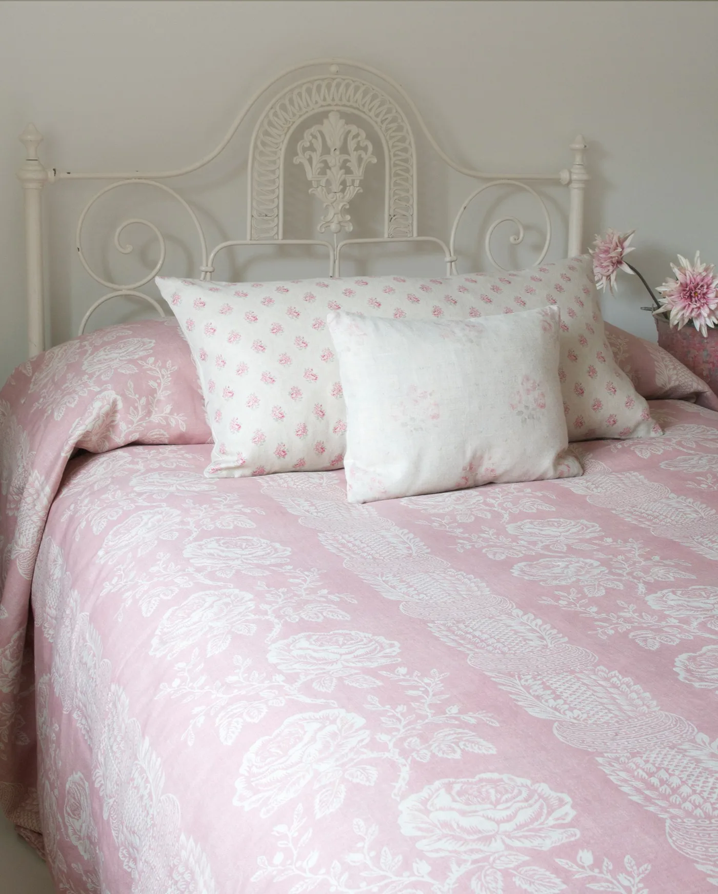 Pale bedroom with an ornate white metal headboard, pink patterned bedding and a pale floral cushion.