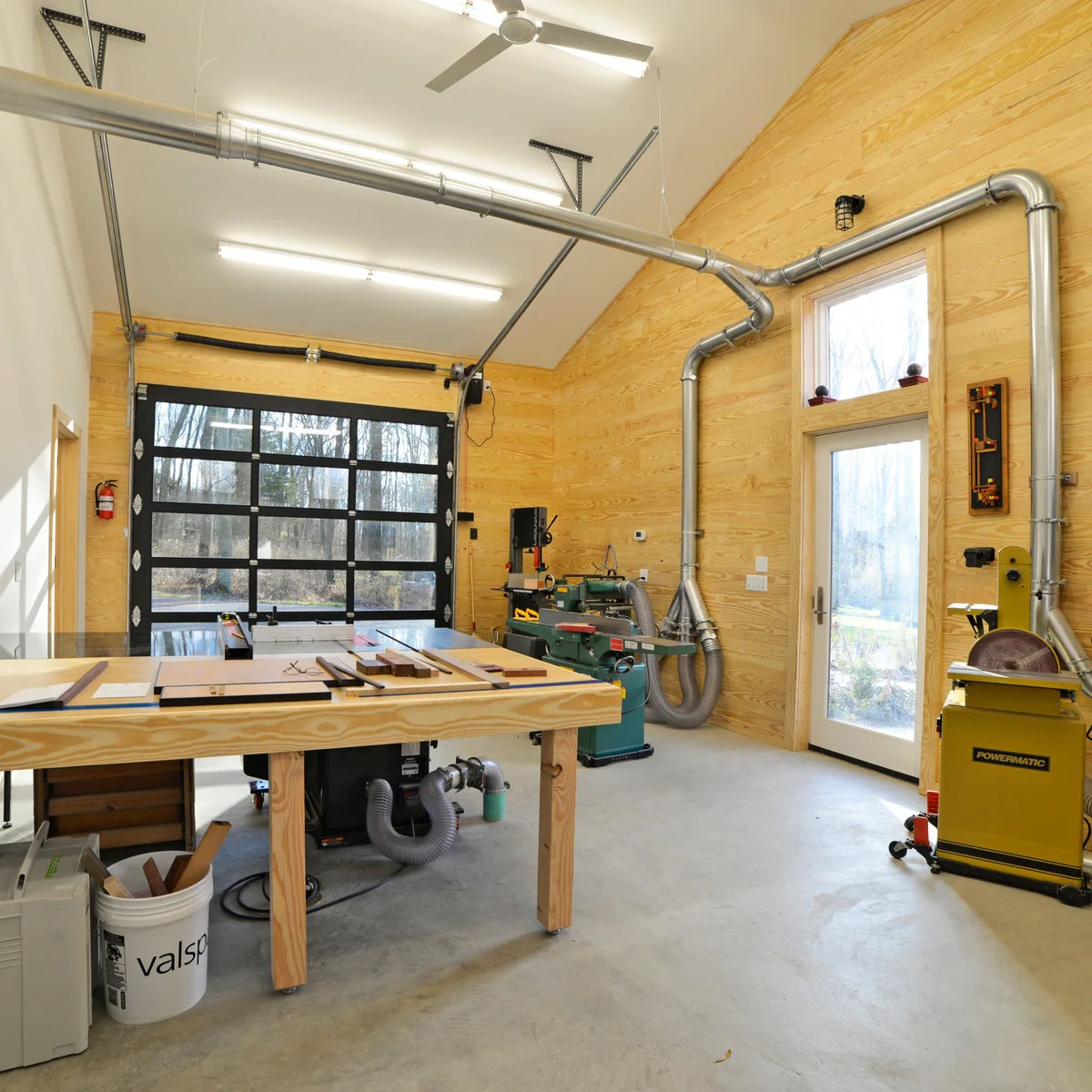Bright wood-lined workshop with a central saw table, other machines along the wall and overhead extraction ducting.