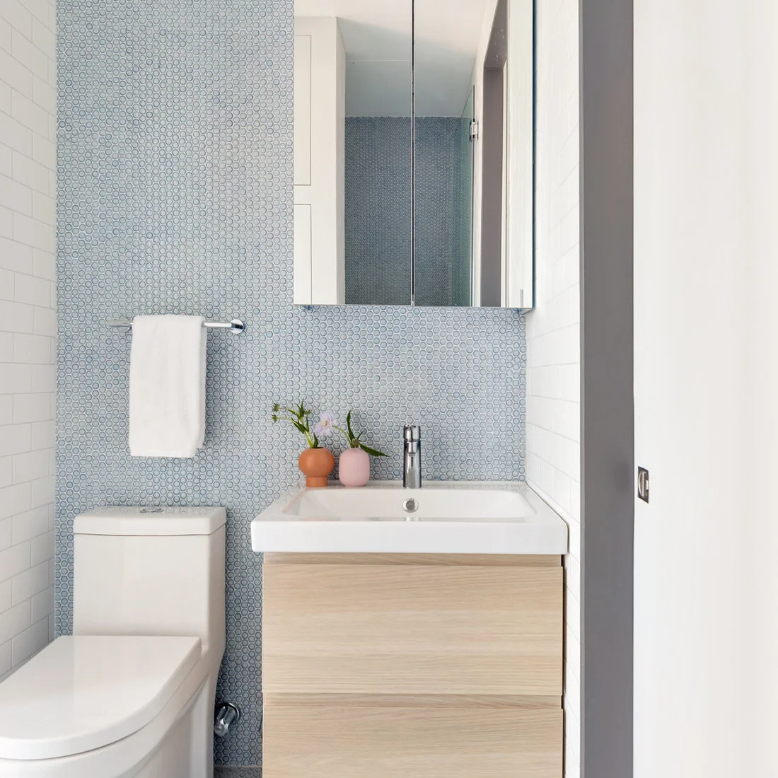 A narrow powder room has pale blue small circular tiles behind a white basin, mirrored cabinet and toilet, with a light wood drawer vanity.