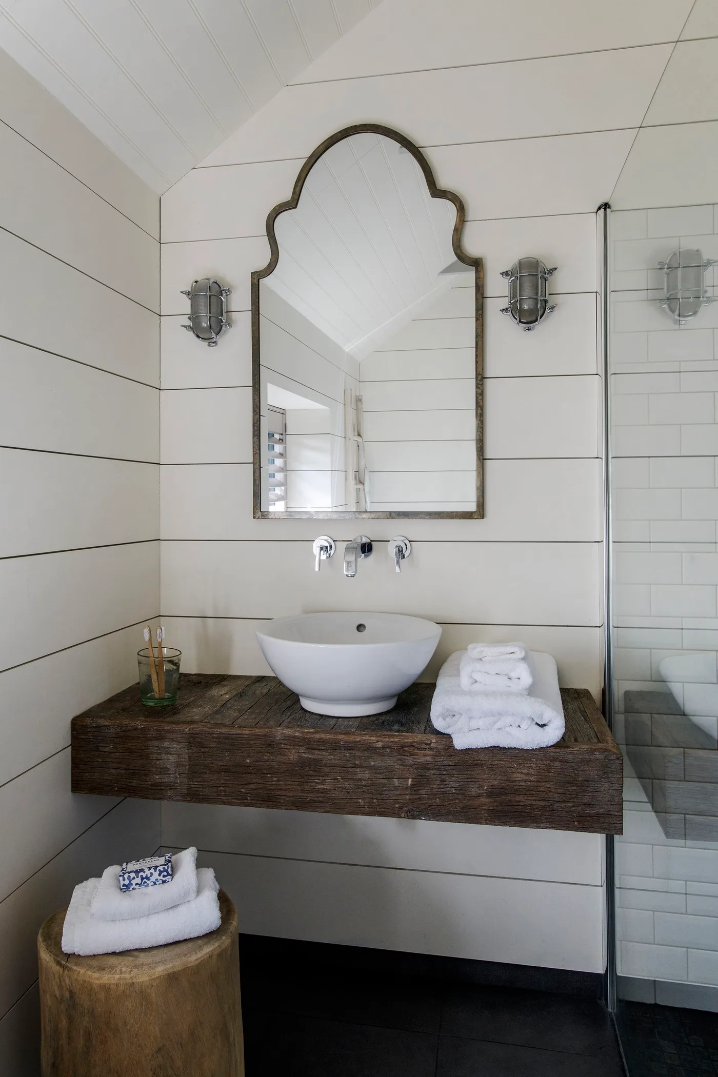 Bathroom with white horizontal wall boards, a wooden basin shelf, white vessel basin, shaped mirror and clear shower screen.
