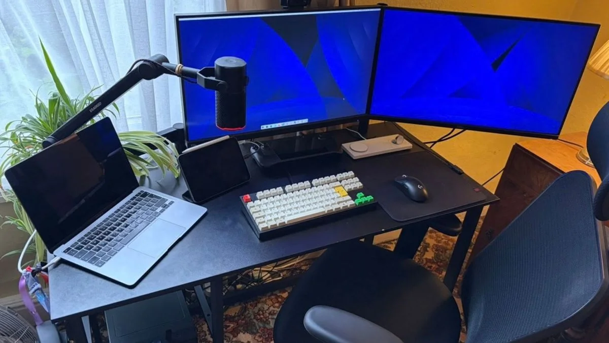 A black desk beside a window with two monitors, a laptop, a compact keyboard, a microphone arm and a padded office chair.