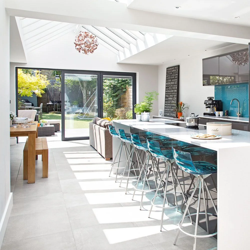 Turquoise stools line a white kitchen counter beside a wooden dining table, glass garden doors and a glazed roof