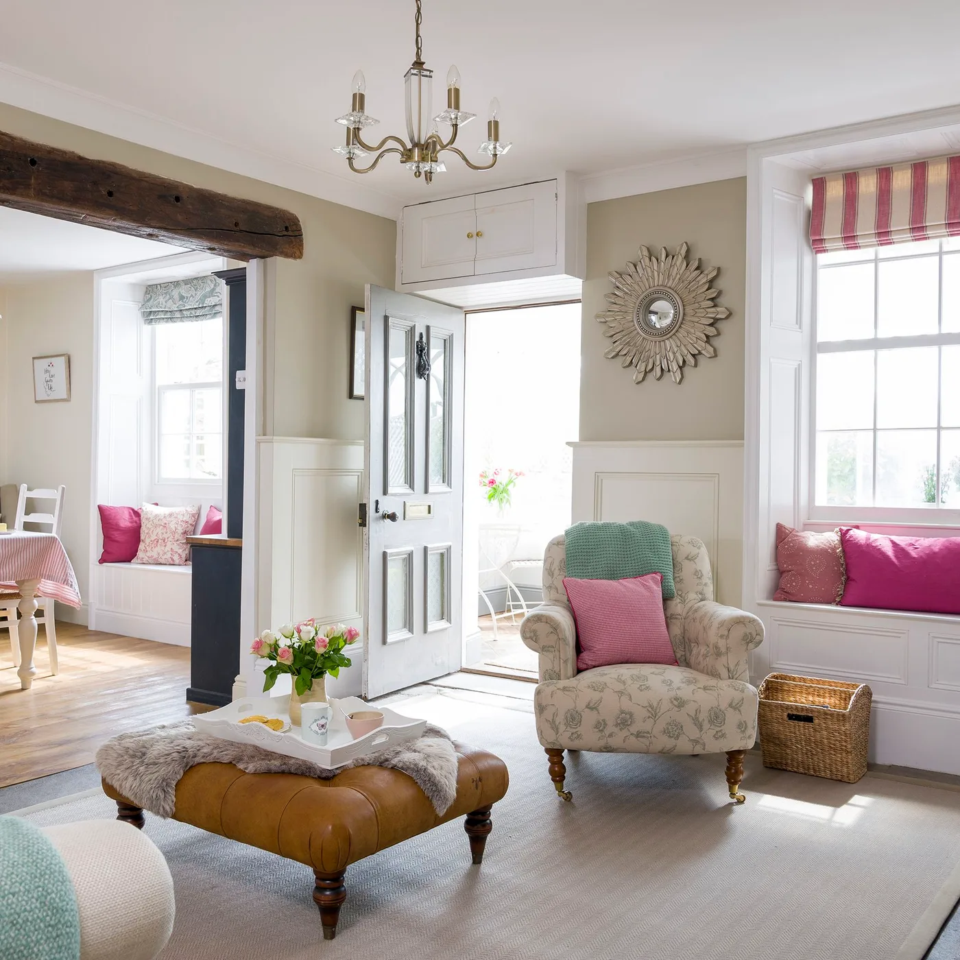 Open front door leading into a cottage sitting room with pale paneling, a cushioned window seat, floral chair and adjoining dining area.