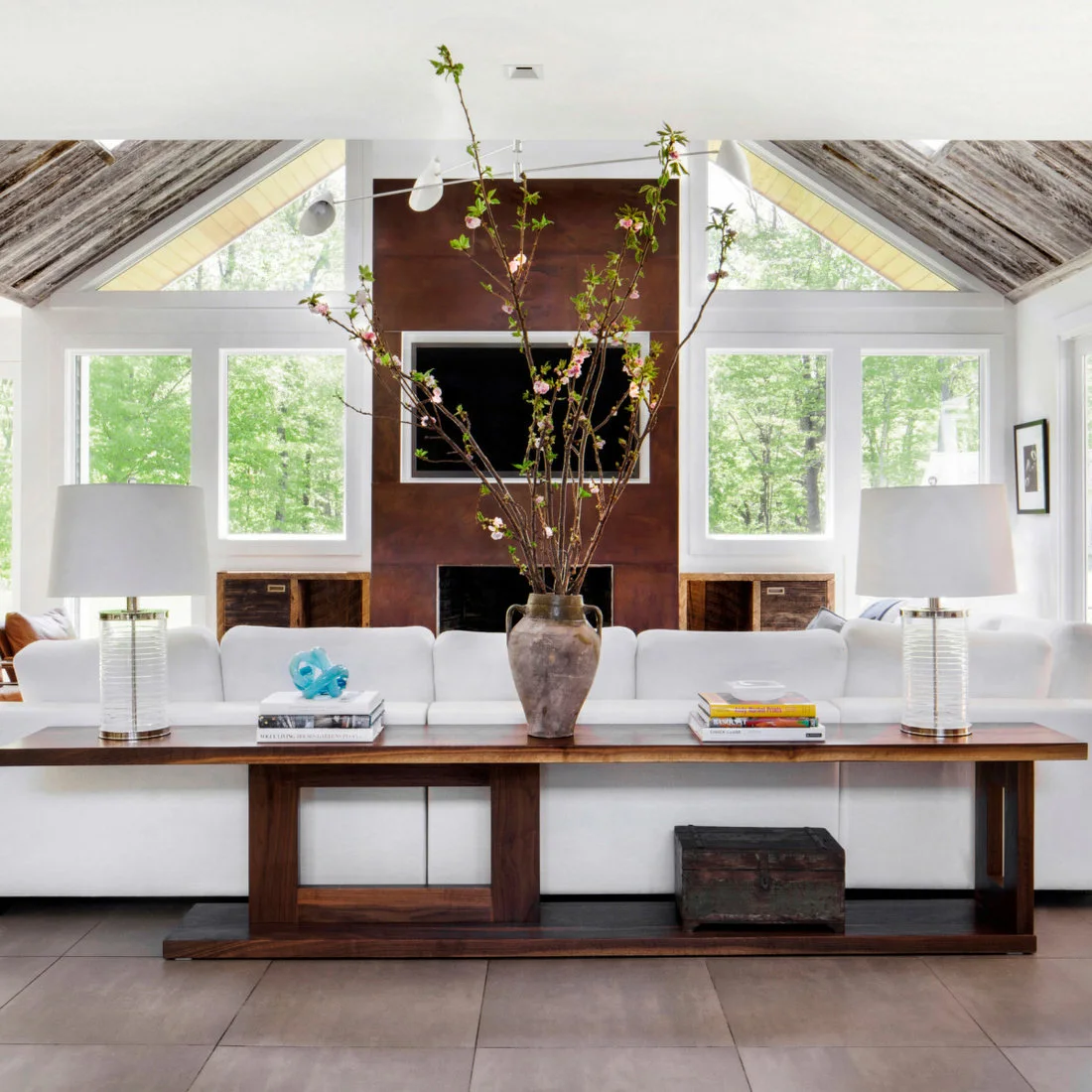 A long timber console behind a white sofa holds two pale lamps, books and a tall branching vase.
