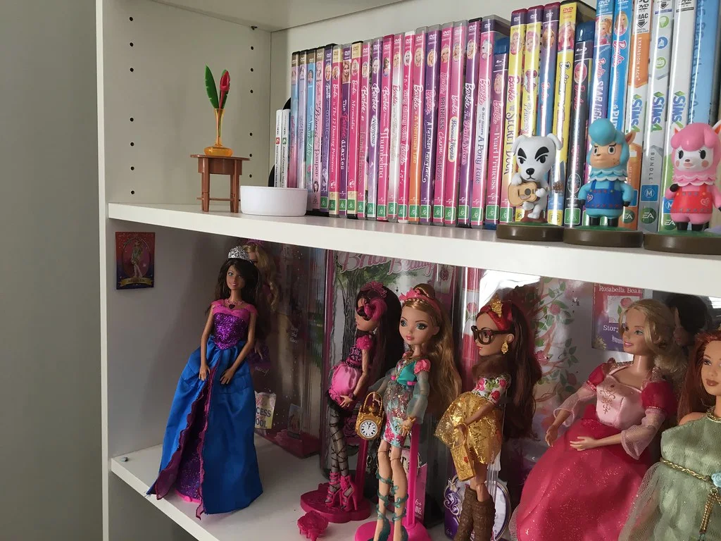 White shelving with boxed and unboxed fashion dolls below a row of Barbie DVDs.