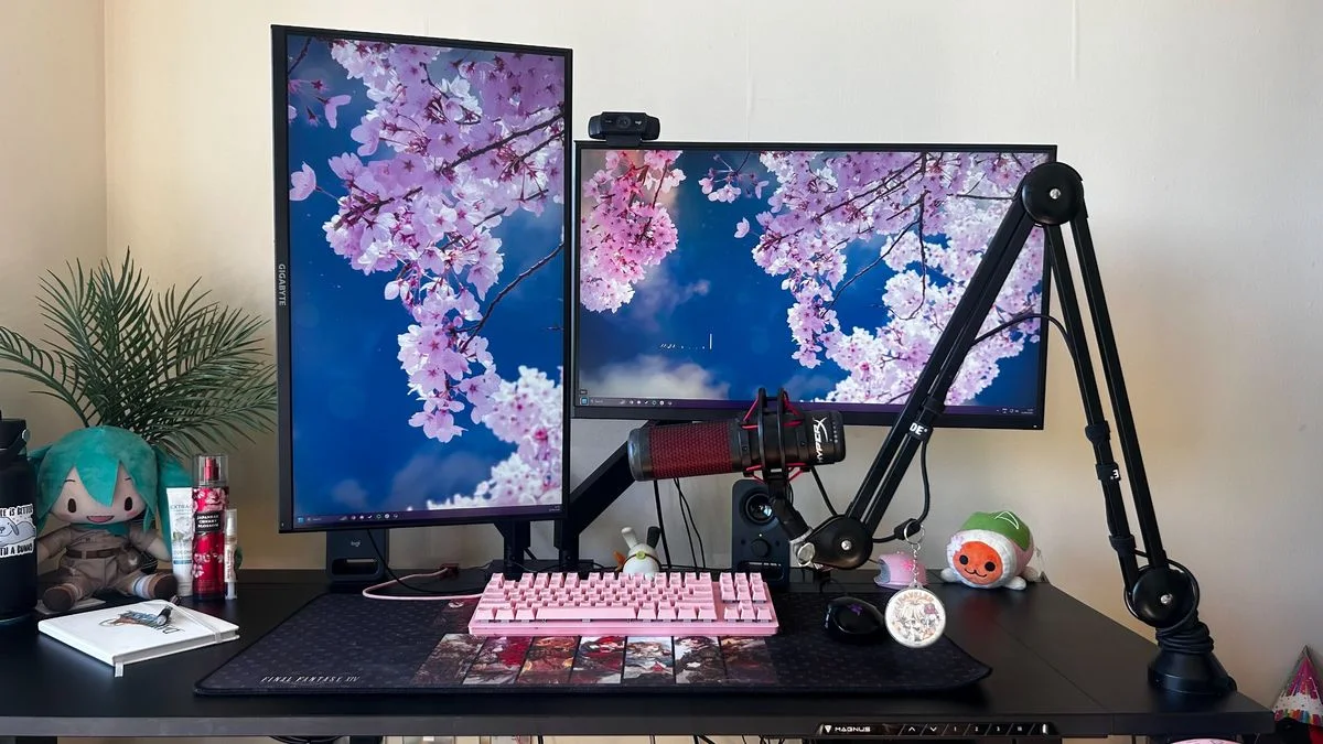 A black desk with two monitors showing pink blossoms, a pink keyboard, a microphone arm and small character toys.
