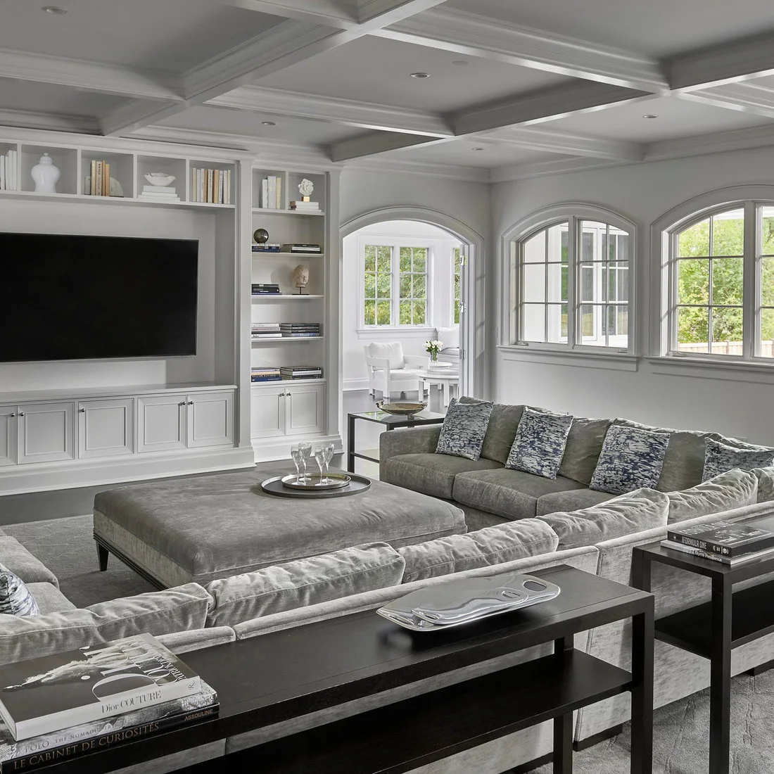 Two dark open-frame consoles with books and a silver tray run behind a gray sectional.