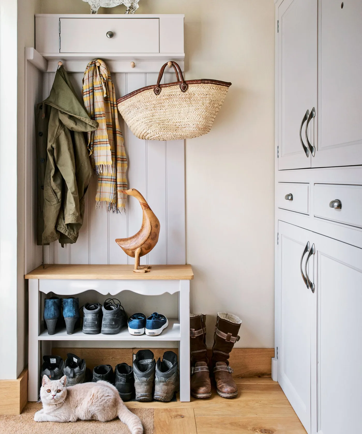 Small pale hall tree with wooden seat, shoe shelves, coat pegs and upper drawer beside taller cupboard.
