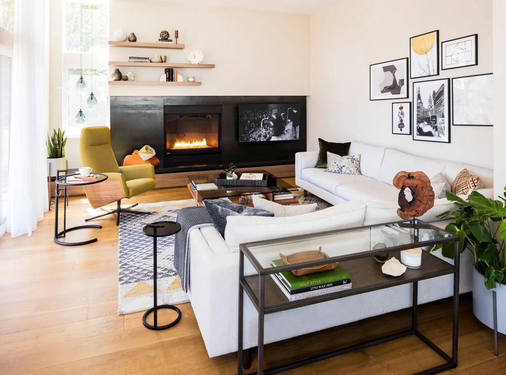 A rectangular fire opening and television sit within a broad black horizontal wall section below wood display shelves beside pale sofas and a yellow chair
