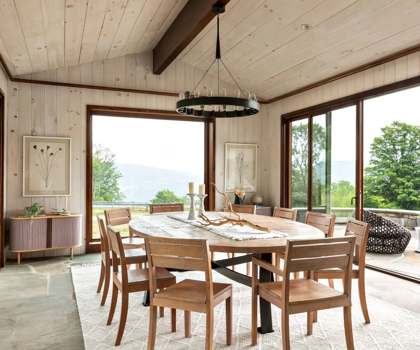 Country dining room with pale wooden wall and ceiling boards, dark roof beam, round table, wooden chairs and broad windows.