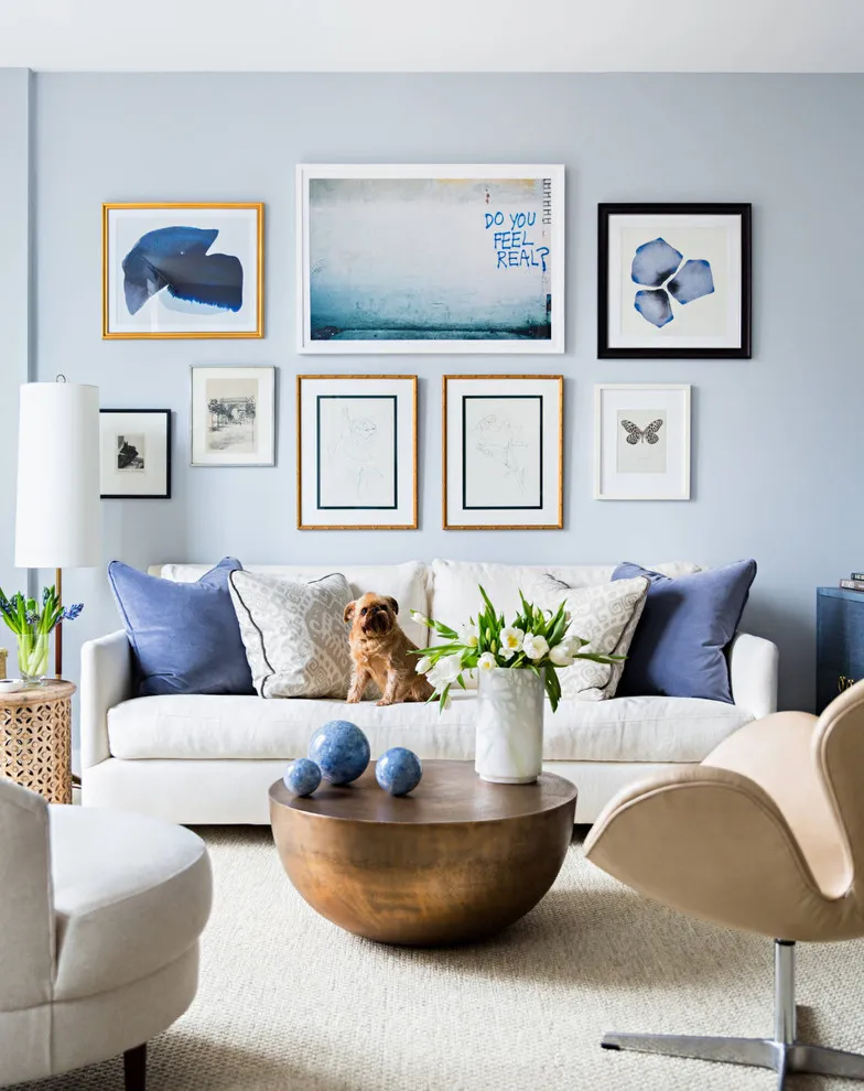 A mixed group of pale and dark frames hangs above a white sofa with blue cushions