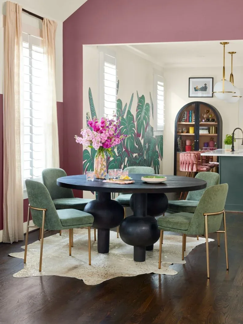 Mauve dining-room wall around a broad opening, black round table, green upholstered chairs, pale curtains, flowers, and botanical imagery in the adjoining room.