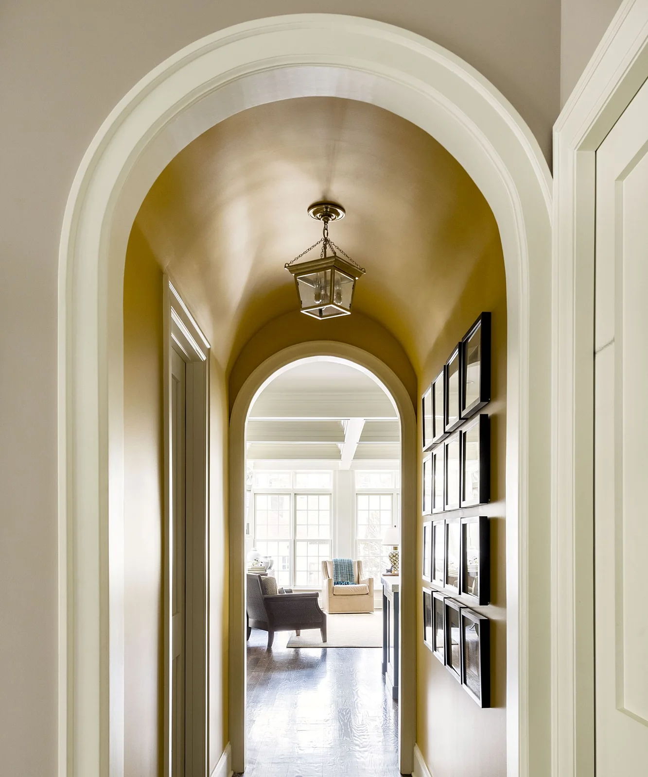 A yellow arched hallway carries rows of small framed pictures beneath a hanging lantern and leads through white trim to another room