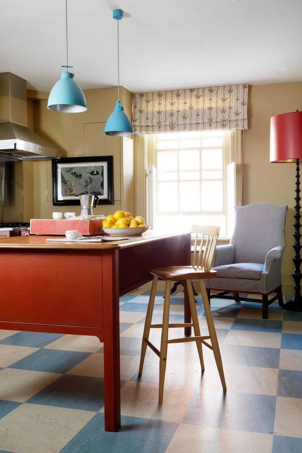 Red kitchen cabinetry and an island stand beneath blue pendants beside a blue-and-pale checked floor, patterned blind and striped chair