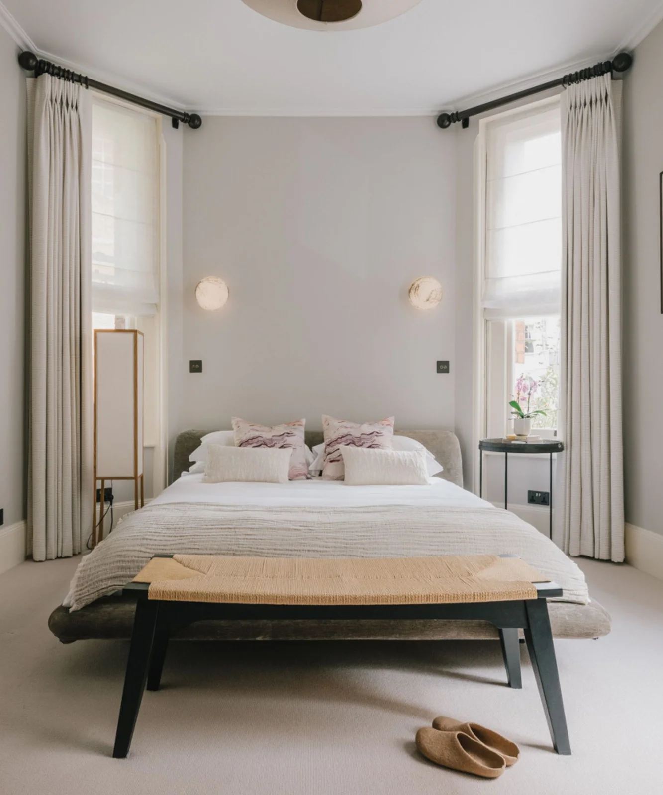 A pale bed stands between tall curtained windows beneath two round wall lights with a narrow woven bench at its foot