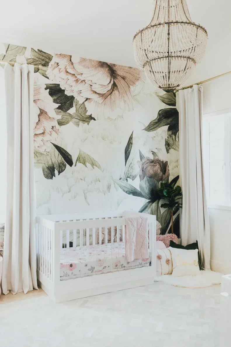 White crib against large floral wallpaper with lighter curtains, decorative pendant and pink staged fabrics.