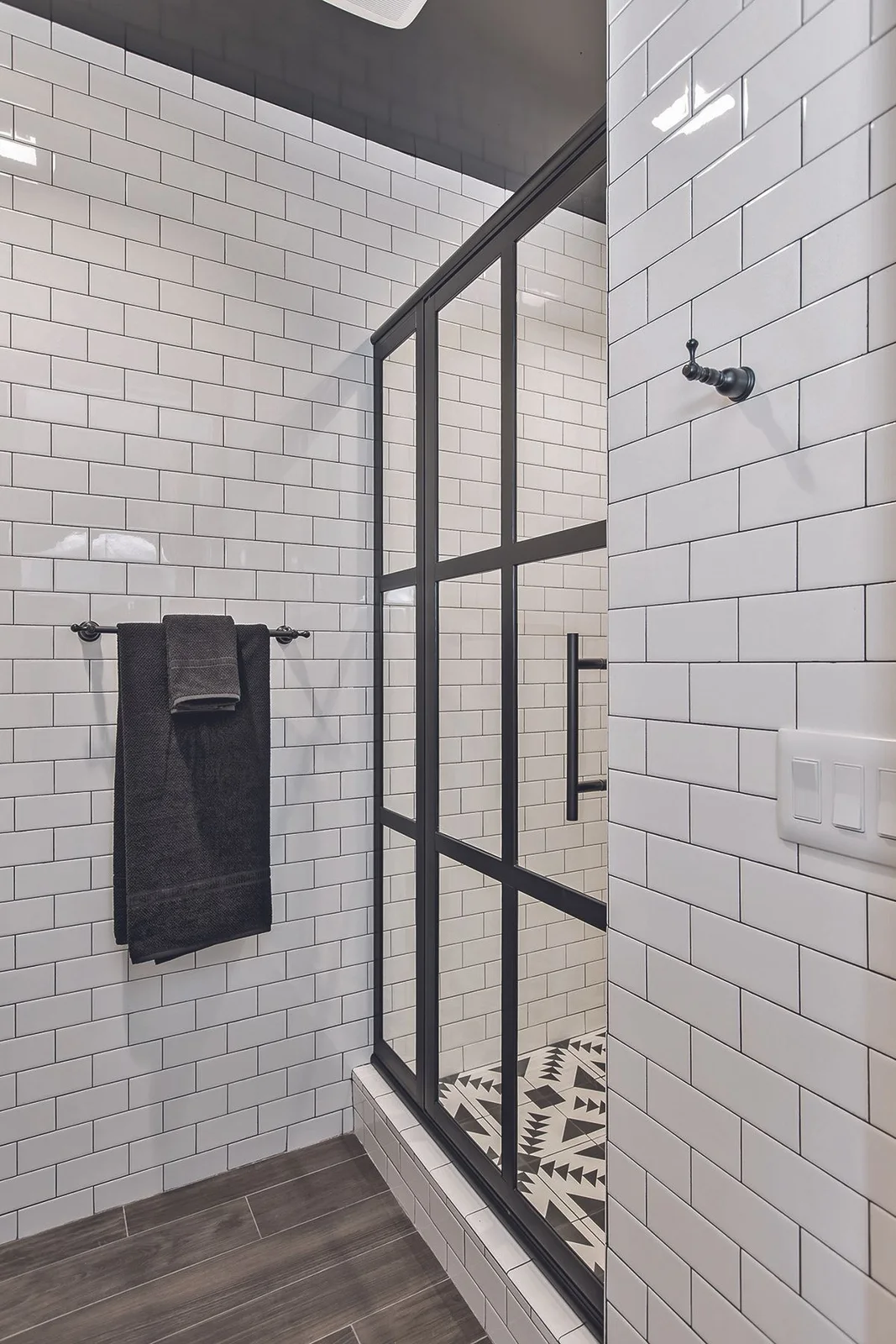 White subway tiles with dark grout surround a black-framed shower screen beside gray towels, a dark upper wall and a warmer wood-colored floor