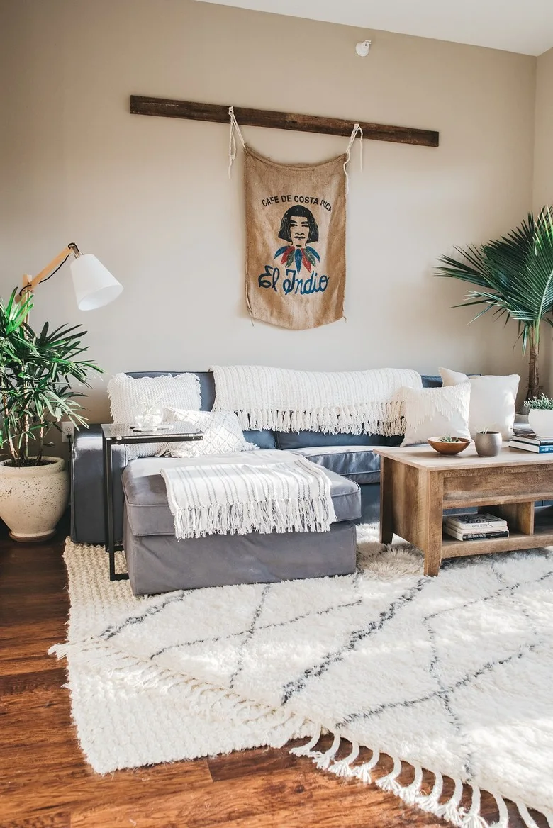 A gray sectional and matching ottoman have pale fringed throws, with a wood table, large plants, a textured pale rug and a fabric wall hanging.