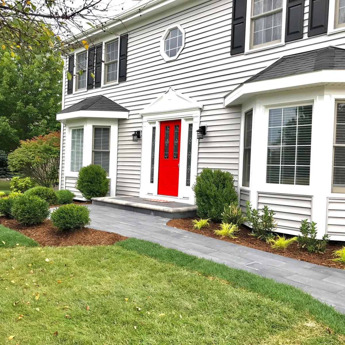 A gray paved front walk beside simple mulched beds, rounded shrubs and a white house with a red entrance door.