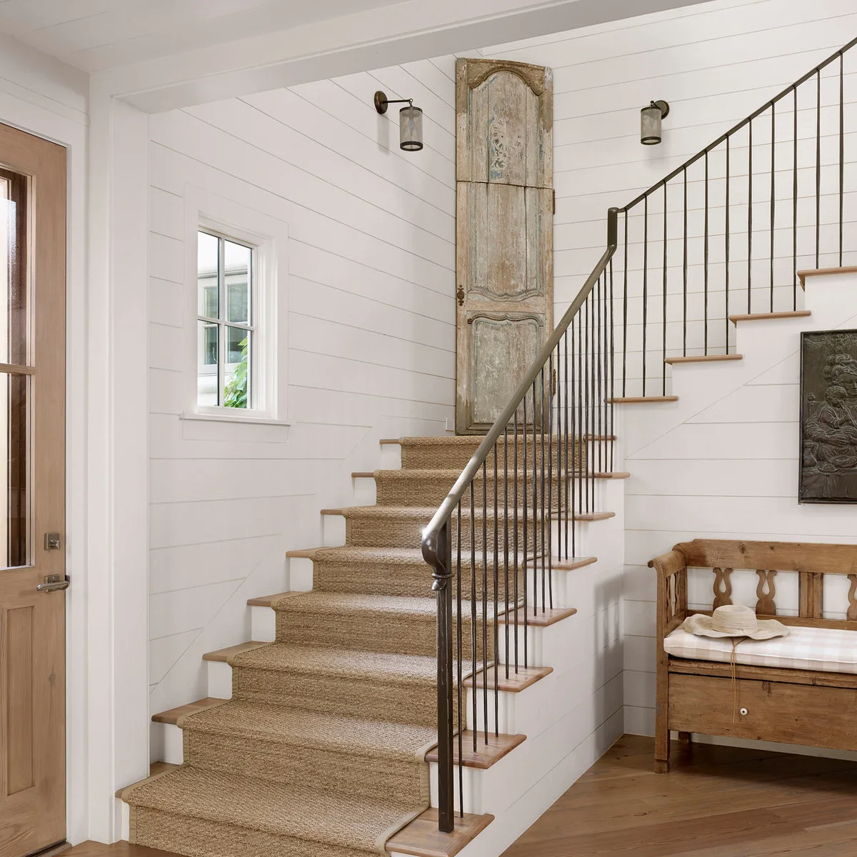 White boarded entry hall with a textured stair runner, slim dark rail and a wood storage bench
