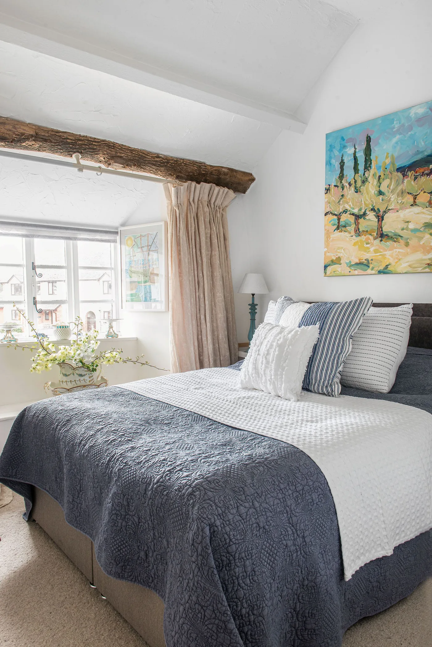 A bedroom has a rough exposed timber above the window, pale curtains, blue-gray bedding, striped cushions and bright landscape artwork.