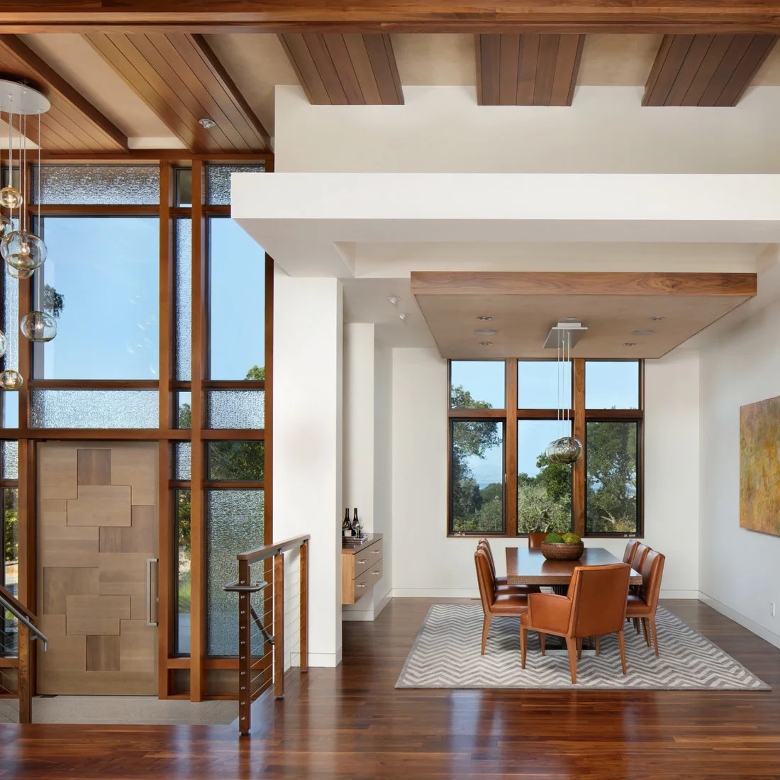 A wood dining table with warm brown padded chairs sits on a pale zigzag rug beneath globe pendants beside wood ceiling beams and a broad window