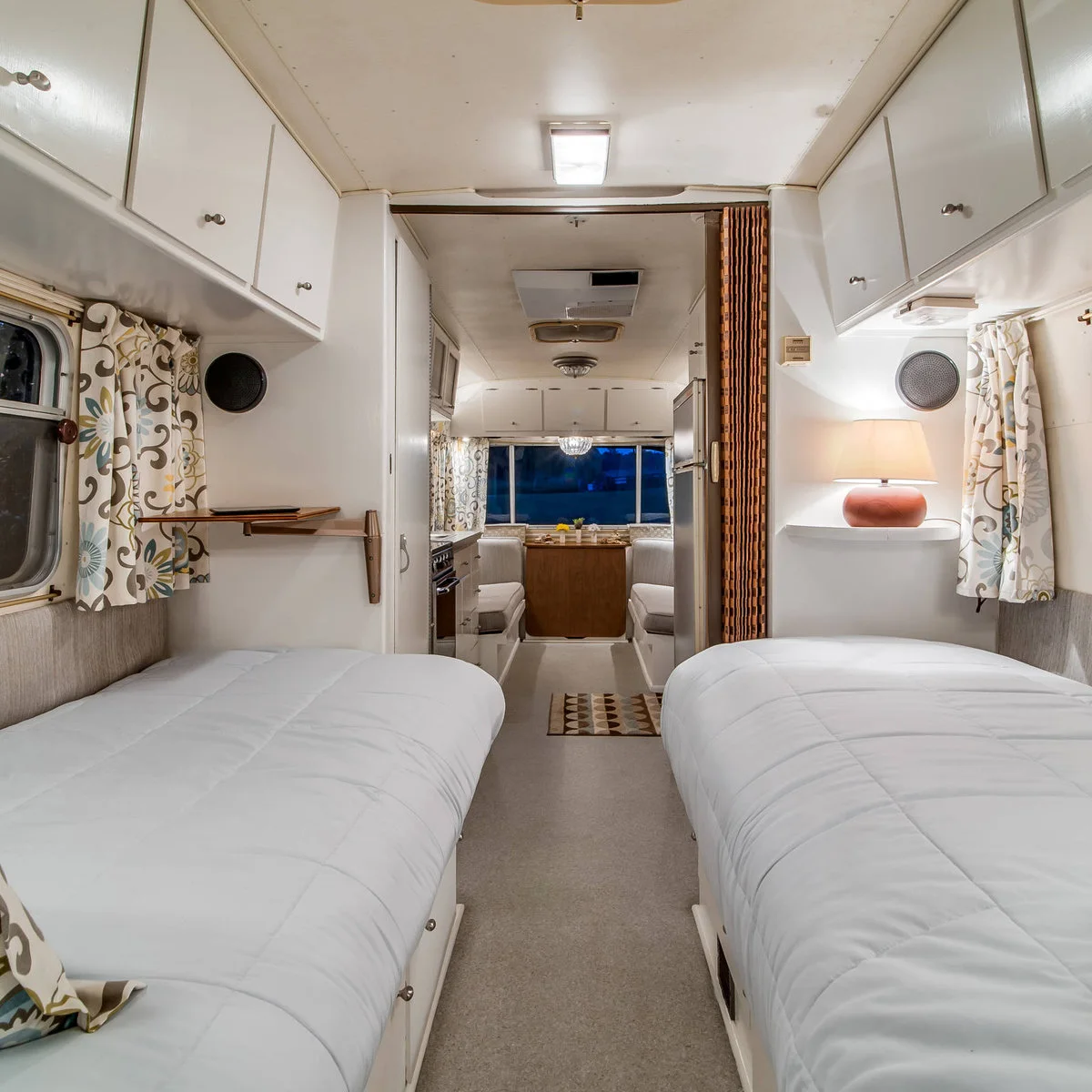 White camper interior with twin beds, overhead cabinets and a central aisle leading toward the dining area.