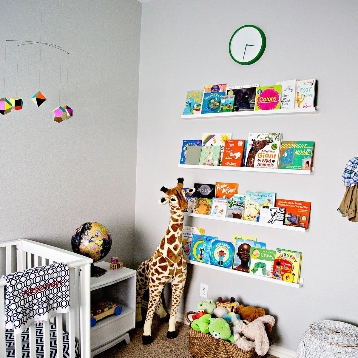 White nursery wall with four face-out book ledges, a green clock and a tall toy giraffe