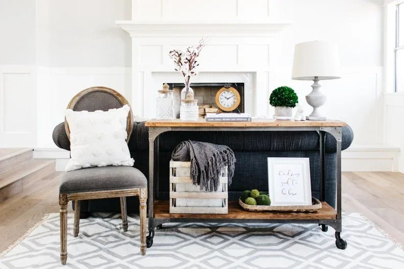 A timber and metal sofa console carries a lamp and glass jars above a crate of throws and framed artwork.