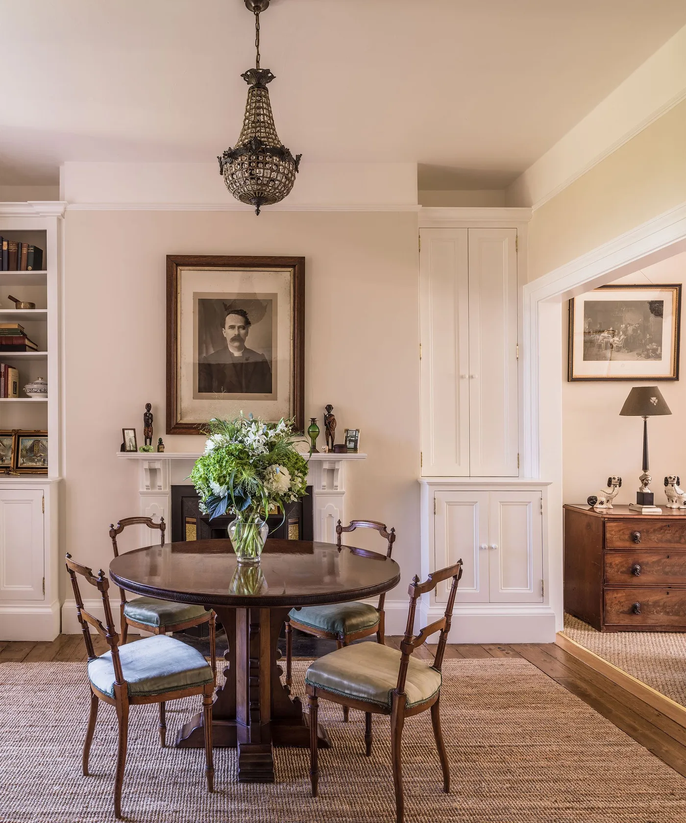 Country dining room with round wooden table, curved chairs, pale fitted cupboards, framed artwork and a fireplace beside an adjoining room.