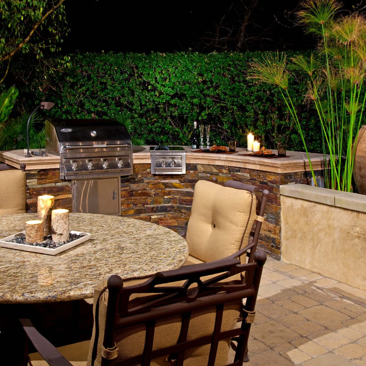 Evening outdoor kitchen with angled counter, metal grill, separate burner and round dining table with cushioned chairs